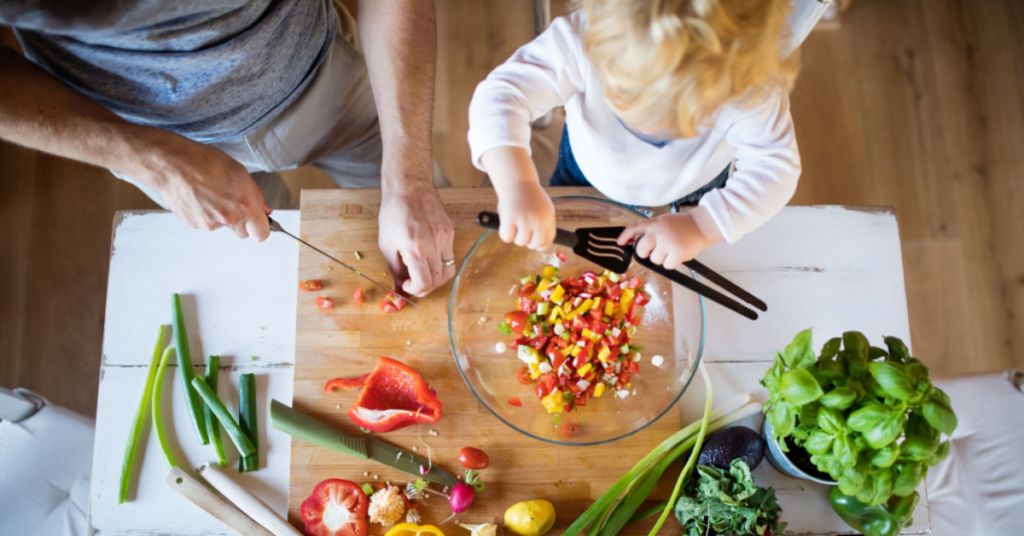 Criança e pai se divertindo na cozinha enquanto preparam uma receita colorida e saudável com alimentos orgânicos.